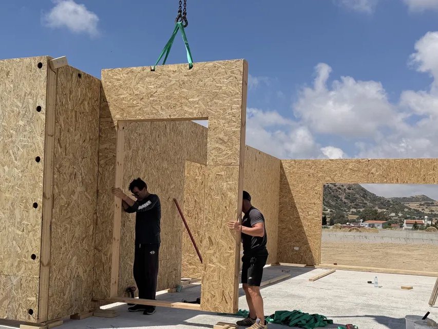 Στήσιμο ξύλινης κατοικίας: ο γερανός κατεβάζει έτοιμο πάνελ τοίχου πάνω στην πλάκα.